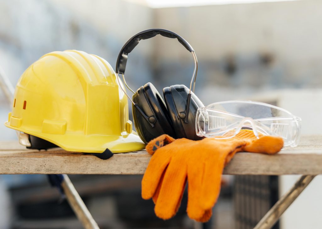 Das Bild zeigt eine Ablage auf einer Baustelle. Darauf liegen ein gelber Schutzhelm, ein paar Schallschutzkopfhörer und ein paar orangefarbene Handschuhe.